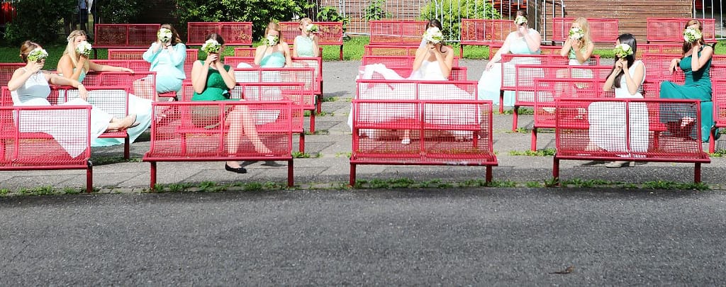 Hochzeitsgesellschaft auf roten Bänken hält sich Blumensträuße vors Gesicht
