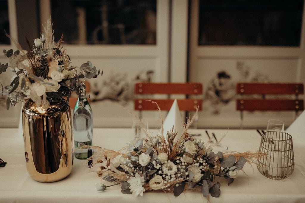 Blumenschmuck aus Trockenblumen in goldener Vase neben Tischgesteck