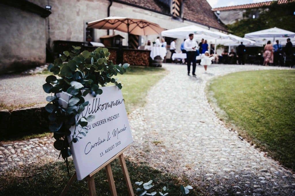 Willkommensschild mit Blumenschmuck, im Hintergrund Hochzeitsgäste
