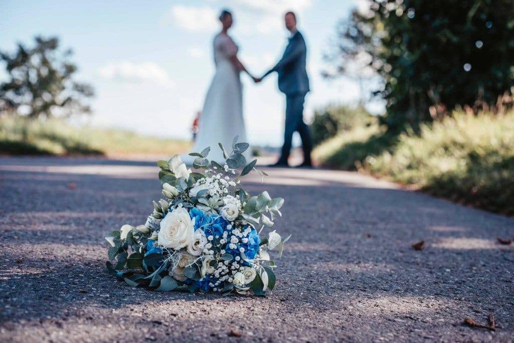 Blau-weißer Brautstrauß auf Straße, Brautpaar Händchen haltend im Hintergrund